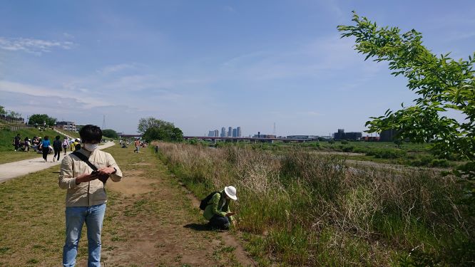 In the foreground of this photo, two people are surrounded by wildlife, one person appears to be looking at their phone and the other is crouched down in tall grass. A city with several high-rise buildings is in the distance behind them.